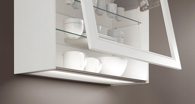 Nowoczesna biała szafka kuchenna prezentująca zorganizowane szkło i naczynia z oświetleniem podszafkowym podkreślającym czysty i minimalistyczny design estetyczny.