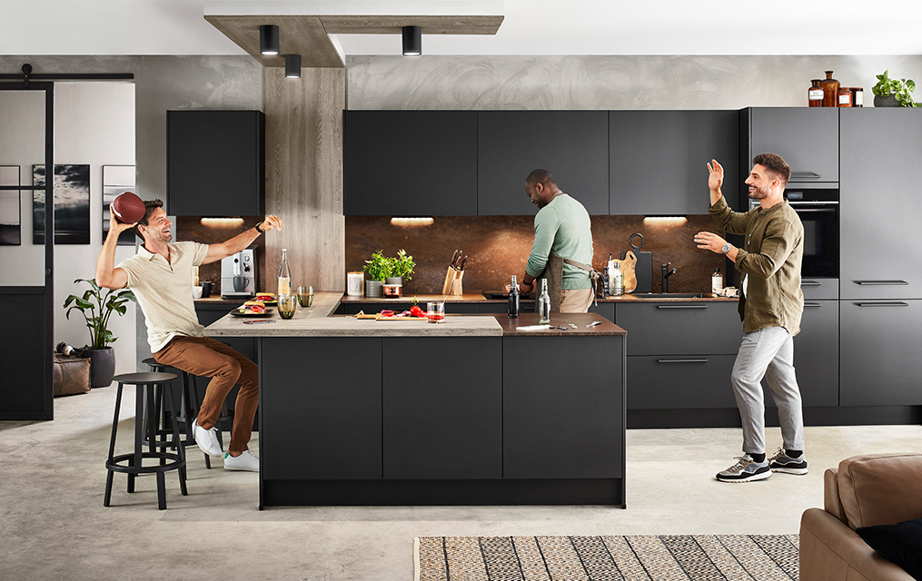 Trzech mężczyzn bawi się podczas swobodnego spotkania w nowoczesnej kuchni z eleganckimi czarnymi szafkami, przygotowując jedzenie i dzieląc się śmiechem.