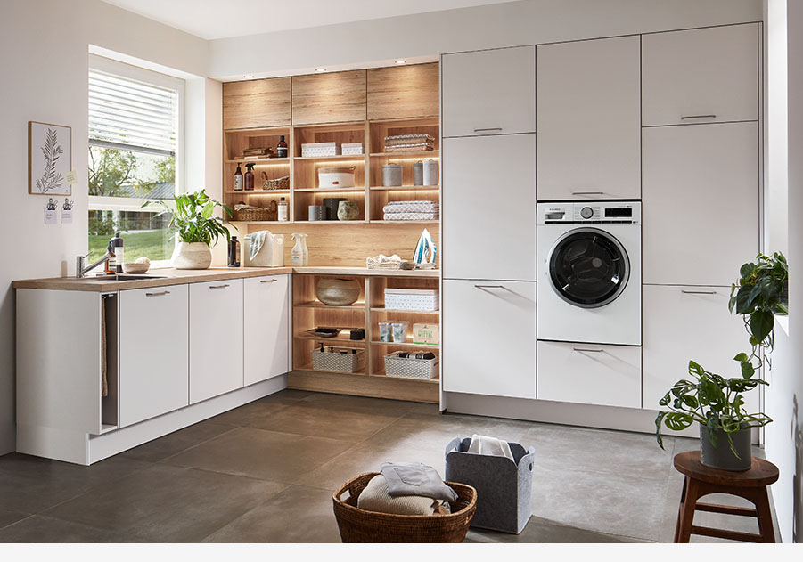 Cocina contemporánea con gabinetes blancos, estantes de madera, electrodomésticos integrados y abundante luz natural creando un espacio acogedor y elegante.