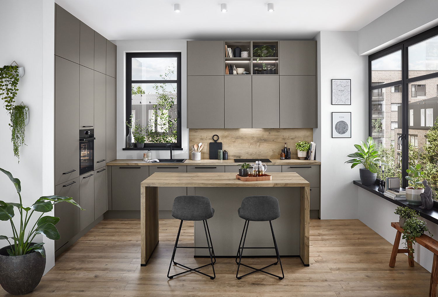 Moderne Küchengestaltung mit eleganten grauen Schränken, Holzakzenten, integrierten Geräten und einer zentralen Insel mit Hockern, beleuchtet von natürlichem Licht durch große Fenster.