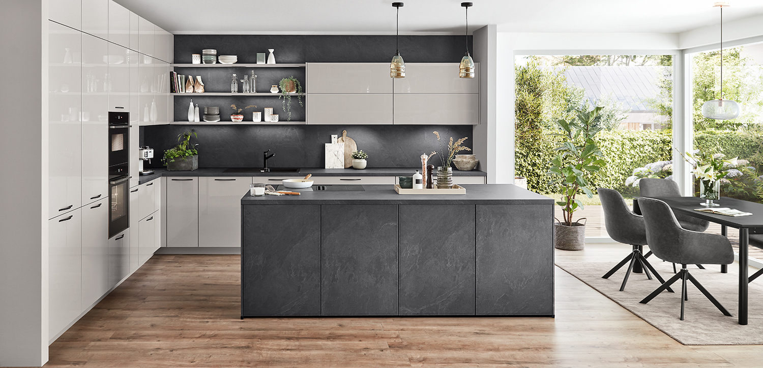 Modern kitchen interior featuring a central island, sleek white cupboards, stainless steel appliances, and pendant lights, with a view of greenery through large windows.