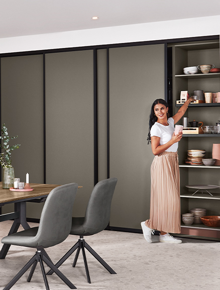 Współczesna jadalnia z uśmiechniętą kobietą obok otwartej półki, nowoczesnymi meblami i minimalistyczną kolorystyką.