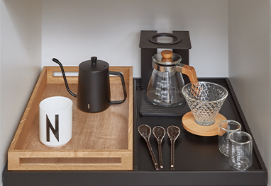 Elegantně uspořádaná kávová stanice s černým konvíkem, mlýnkem, skleničkami a jedinečnými lžičkami na elegantní dřevěné podložce, předvádějící moderní design kuchyňských potřeb.