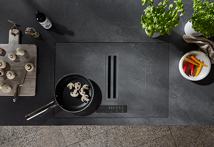 Entorno de cocina moderna con una elegante placa de inducción negra con verduras frescas e ingredientes de cocina dispuestos ordenadamente en una encimera oscura.