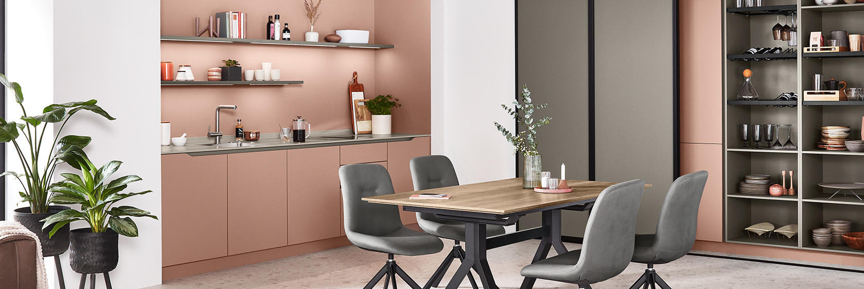 Modern kitchen interior featuring sleek salmon-colored cabinetry, open shelving with books and decor, and a cozy dining area with grey upholstered chairs.