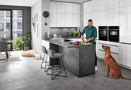 A modern kitchen with clean lines, featuring a person preparing food on a sleek island countertop while a dog watches attentively.