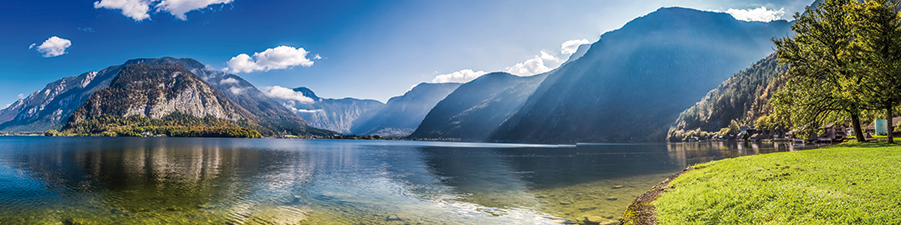 Panoramic view of a tranquil mountain lake with clear waters reflecting the surrounding peaks, under a blue sky with scattered clouds.