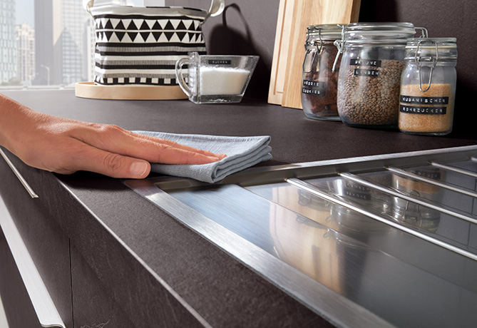 Een hand van een persoon die een moderne keukenwerkblad naast een keramische kookplaat afveegt, met op de achtergrond opbergpotten en een koffiekopje.