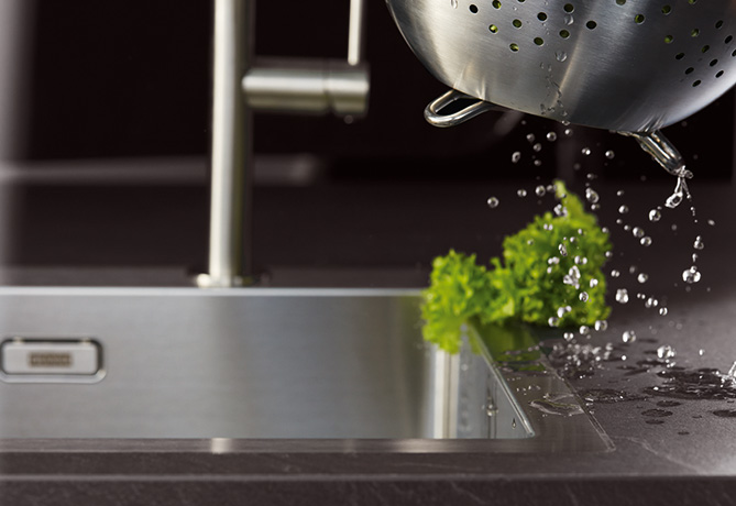 Modern kitchen sink with stainless steel faucet, freshly washed lettuce in a colander, water droplets in motion, and a sleek dark countertop.