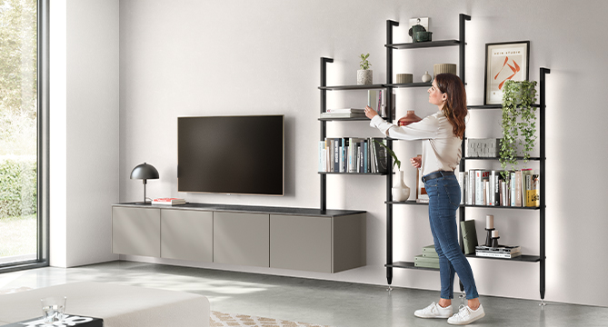 Moderní obývací pokoj s stylovou zábavní jednotkou, regálovým systémem a ženou, která uspořádává předměty na policích, odrážející uklizený, moderní design.