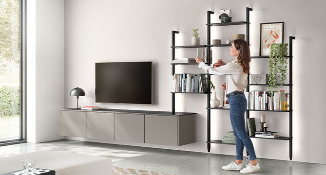 Soggiorno moderno con un elegante mobile porta TV, sistema di scaffalature e una donna che dispone oggetti sugli scaffali, riflettendo un design ordinato e contemporaneo.