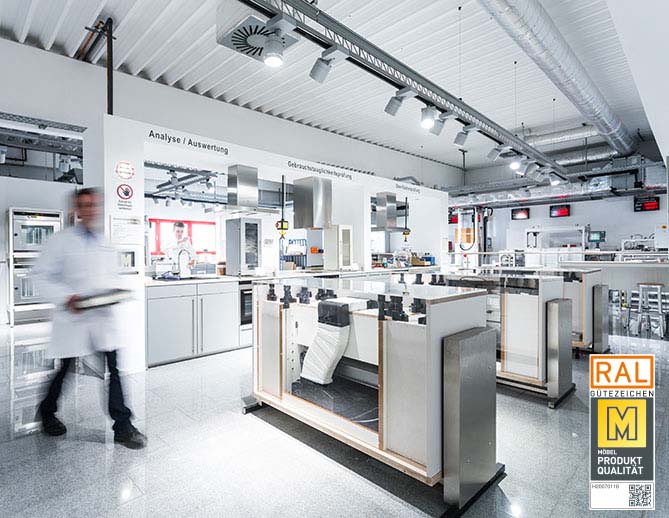 Un laboratorio moderno e pulito con postazioni di lavoro in acciaio inossidabile, attrezzature tecniche e la figura sfocata di un professionista in camice in movimento.