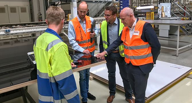 Industrial professionals in high-visibility vests engage in a discussion on a factory floor, examining a product or process with focused attention.