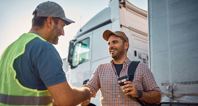 Dwóch uśmiechniętych mężczyzn podaje sobie ręce na rampie załadunkowej dla ciężarówek, z których jeden nosi kamizelkę odblaskową, symbolizując udaną dostawę lub partnerstwo w logistyce.