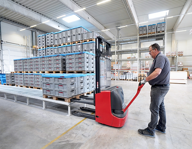 Een werknemer manoeuvreert een palletwagen beladen met kratten in een moderne, goed georganiseerde magazijn, waar efficiënte logistiek en voorraadbeheer worden getoond.