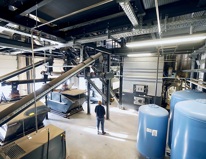 Instalación industrial moderna con maquinaria grande, sistemas de transporte y tanques de almacenamiento, supervisada por un técnico que camina por el suelo limpio y organizado.