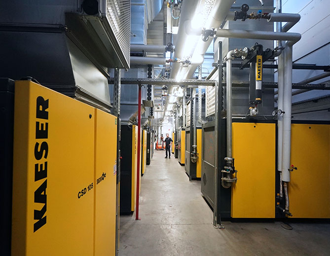 Een persoon loopt tussen rijen grote gele en zwarte industriële Kaeser compressoren in een goed georganiseerde en verlichte faciliteit met overheadleidingen.
