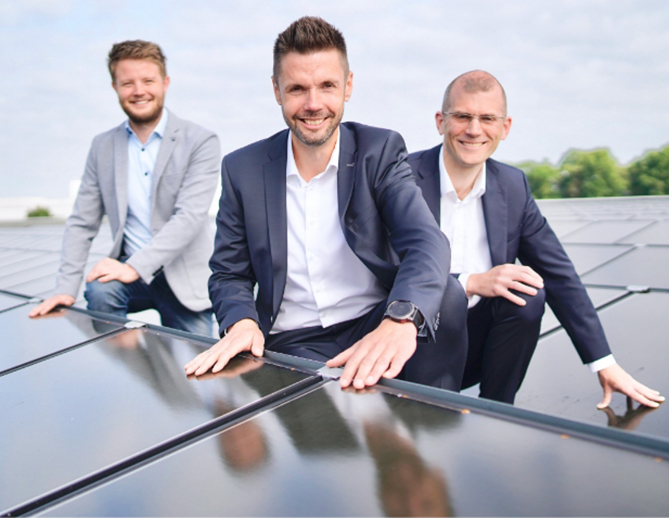 Three professional men in suits smiling on a rooftop with solar panels, symbolizing teamwork and commitment to sustainable energy solutions.