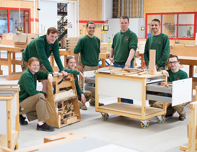Een team van lachende werknemers in groene shirts poseert trots met hun houtbewerkingsproject in een goed uitgeruste werkplaats.