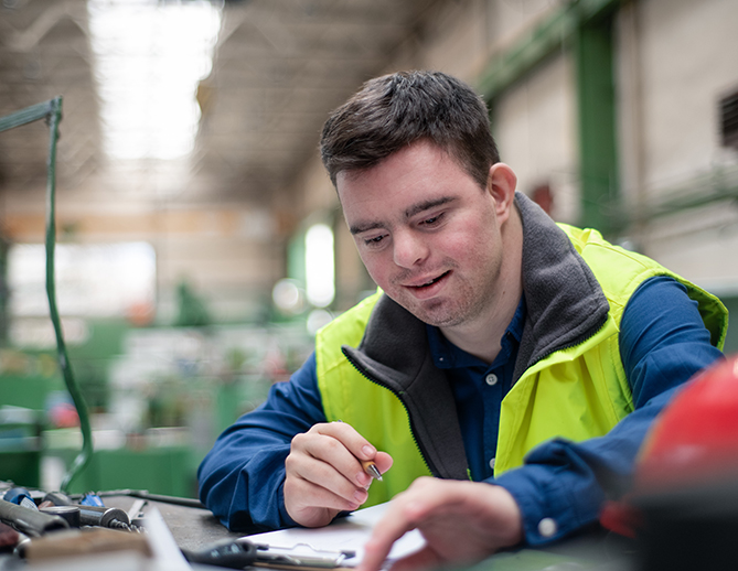 Un travailleur concentré dans un gilet de haute visibilité examine attentivement une pièce dans un environnement industriel animé, témoignant de son dévouement et de son souci du détail.