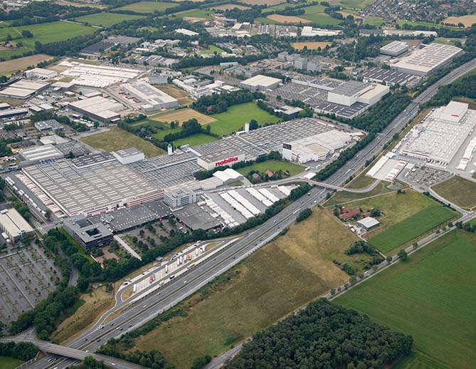 Aerial view of an industrial area with multiple large warehouses and factories, near a highway surrounded by patches of green fields.