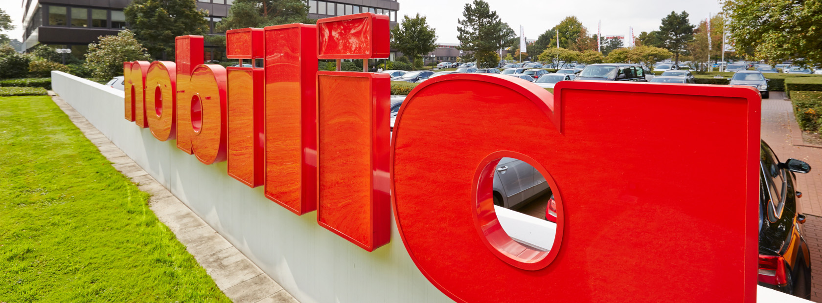 Trójwymiarowe czerwone litery tworzące słowo zainstalowane na zewnątrz, z bujną zielenią i nowoczesnym budynkiem w tle.
