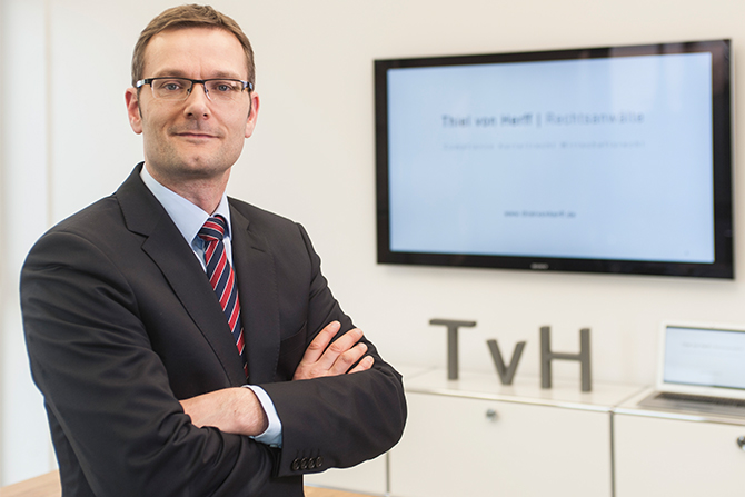 Professional man in a suit with glasses stands confidently in an office environment, with a presentation screen and company acronym in the background.