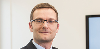 Professional man in business attire with glasses poses confidently in a modern office setting, embodying leadership and expertise.