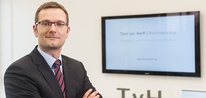 Ein professioneller Mann in einem Anzug mit Brille steht selbstbewusst in einer Büroumgebung, mit einem Monitor im Hintergrund, auf dem Text angezeigt wird.